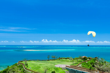 沖縄本島の知念岬公園から望むエメラルドグリーンの海の上空を遊覧するパラグライダー - obrazy, fototapety, plakaty