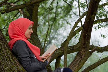 Cute muslim student reading a book while sitting on the tree branch in the park. Modern and fashionable female model representing islam and knowladge, education and stressfree. Arabic culture