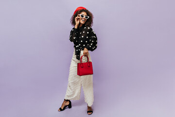 Full length photo of modern woman with curly hairstyle and red beret in black polka dot blouse, wide light pants and dark heels smiling.