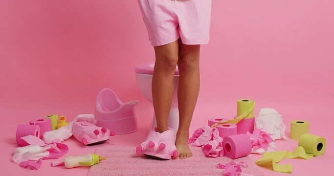 Unrecognizable woman comes in restroom sits on toilet bowl takes shorts and panties off reveals herself wears slipper on one foot feels relaxed poses against pink background has prostate problem