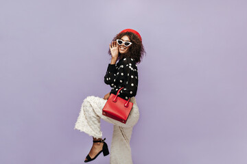 Happy stylish lady with dark short hairstyle and modern sunglasses in red beret, polka dot blouse and trendy pants holding bright handbag.