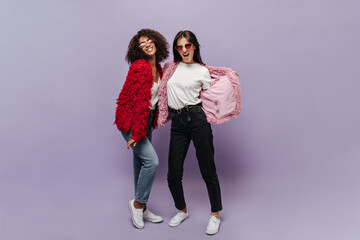 Attractive two young women with brunette hairstyle and red sunglasses in fluffy warm bright sweaters and white sneakers looking into camera.