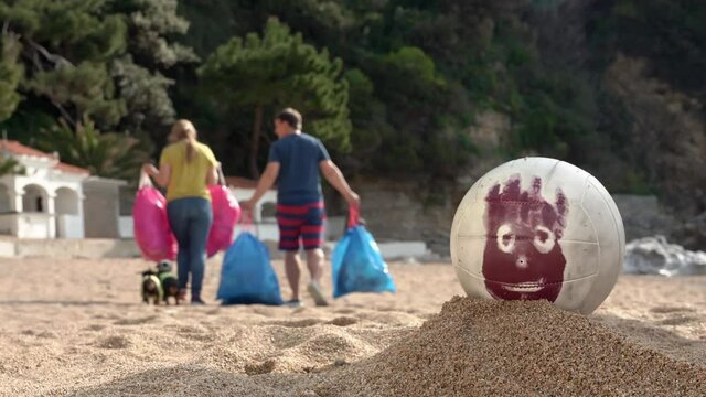 Young man and woman carry away packages, and dachshund dogs run after them. Volunteers have removed garbage on beach and are going to send it to recycling center. Ball with painted face rolled away.