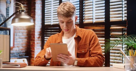 Young handsome student guy sitting alone at home and spending free time chatting with friends on social networks using tablet.