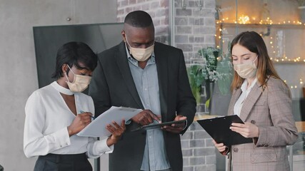 Multiethnic business team of different ages multiracial group of managers workers stands in company office wearing medical masks on faces working during pandemic discussing data in documents papers - Powered by Adobe