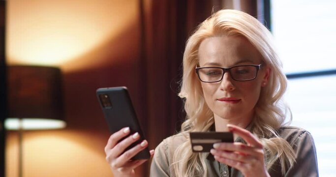 Online store. Happy woman with long blonde hair doing shopping via internet while using smartphone and credit card. Young woman rejoicing of something while sitting at the office. Shopping concept