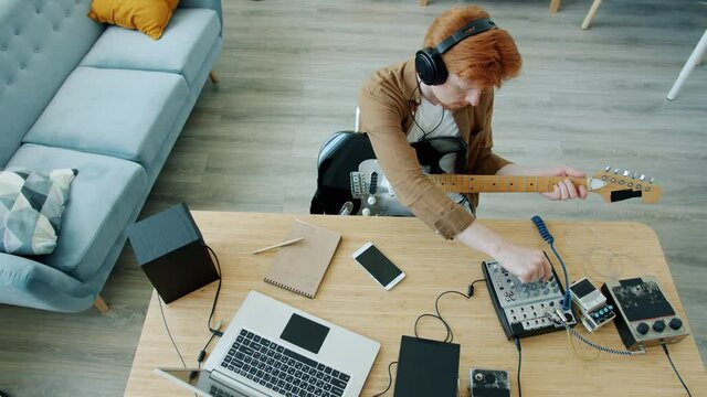 High angle view of ginger-haired man playing guitar and using laptop and sound mixer in apartment. Modern technology and creative people concept.