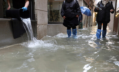 worker empties the water from her shop with a bucket and other people with waterproof gaiters as...