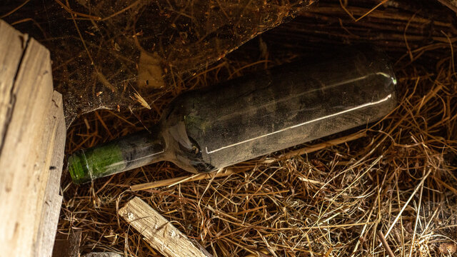 An Abandoned, Forgotten Bottle Of Wine In Dust, Dirt And Hay Among Pieces Of Wood And Firewood Lies In The Corner In The Shade. An Unexpected Find.