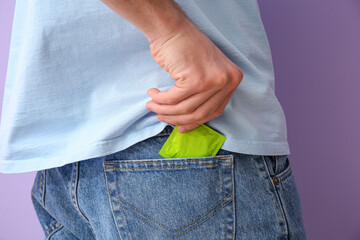 Man putting condom into pocket of jeans on color background, closeup
