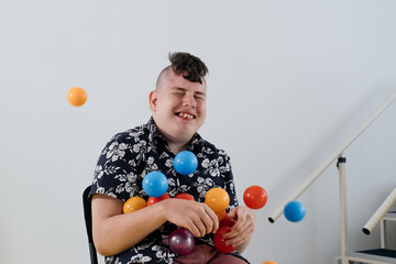 Happy teenage child over white wall in rehabilitation center. Boy with cerebral palsy smiling, playing with balls. Rehabilitation and inclusion kindergarten, day care. Disabled kid, positive emotions 