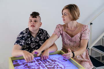 Children with disability getting sensory activity Sand tray, sand table for animation, cerebral palsy boy playing calming game, training fine motor skills. Rehabilitation center with therapist, mother