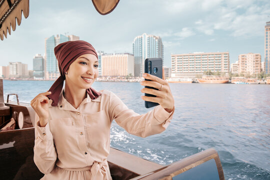 Happy asian woman in a maroon turban takes a selfie on her smartphone while cruising on a traditional Abra Dhow boat on Dubai Creek.