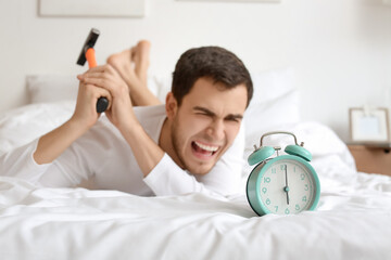 Angry young man with alarm clock and hammer in bedroom