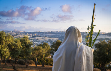 Obraz premium Succot (Feast of Tabernacles) in Jerusalem: Jewish man in a Tallit praying while waving the Four Species, with a view towards the Temple Mount, the Old City and the Mount of Olives