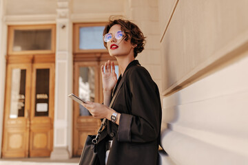Wavy-haired girl with red lips in black outfit posing outside. Trendy woman in glasses with handbag...