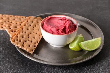 Bowl with tasty beet hummus, lime and crackers on dark background
