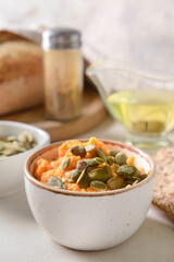 Bowl with tasty carrot hummus, pumpkin seeds and crackers on light background, closeup