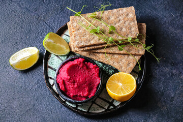 Bowl with tasty beet hummus, lemon, lime and crackers on dark background