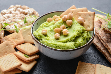 Bowl with tasty green pea hummus, chickpeas and crackers on dark background, closeup