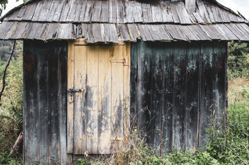 Fototapeta na wymiar An old small Carpathian wooden house, blackened with age, with a traditional wooden tile roof. A yellow grunge door with peeling paint is closed with a rusty iron bolt with hinges made by a blacksmith