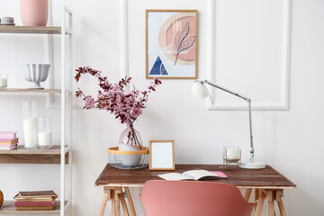 Modern workplace with vase and blossoming branches near light wall