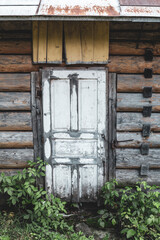 White old weathered door in the wooden wall of an ancient hut, traditional log, paint is peeling. The threshold of an abandoned house is overgrown with bushes. Padlocked on a rusty chai. Vertical.