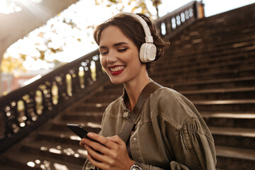 Cheerful woman in olive denim jacket holds phone and smiles outdoors. Brunette lady with red lips in headphone poses on background of stairs..