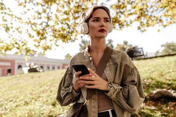 Wonderful lady with short hair in white headphones and olive jacket looking away outdoors. Girl with handbag holds phone outside..