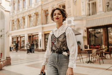Lovely woman with wavy hair in jeans with belt and flower smiling at street. Cool lady in white...