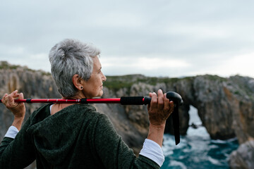 older woman with gray hair crossing a trekking pole between her shoulders and gazing out to sea...