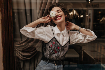 Positive girl with short hair in long sleeve white and black shirt smiling sincerely in cafe. Cheerful lady in jeans holding flower inside..