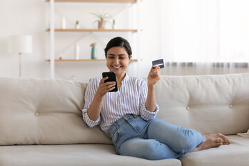 Excited Indian woman paying online by credit card for goods, using smartphone, sitting on couch at home, happy young female satisfied customer browsing internet banking service on device, shopping