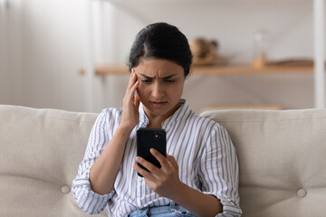 Unhappy Indian millennial woman looking at phone screen, sitting on couch, reading bad news in...