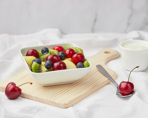 Fresh, healthy, organic fruit salad with cherry, blueberry, kiwi and apple in a white bowl on a cutting board with yogurt and spoon. Closeup shot