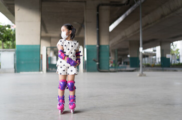 Asian child cute or kid girl exercise playing pink rollerblade or inline skates on sport skating rinks or park and wearing knee to wrist support and elbow pads with white face mask for safety protect