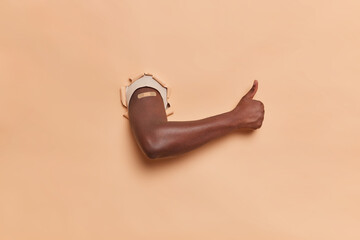 Horizontal shot of dark skinned faceless man shows arm with plaster after vaccination keeps thumb up recommends vaccine against coronavirus breaks through paper background. Antiviral immunization