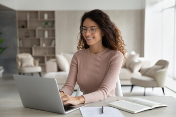 Fototapeta Happy millennial Latino woman in glasses sit at desk at home office look at laptop screen study online. Smiling young Hispanic female work type on computer, consult client customer distant on web. obraz