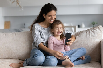 Smiling young Latino mom and biracial teen daughter rest on couch at home have webcam virtual event on smartphone. Happy Hispanic mother and ethnic girl child talk speak on video call on cellphone.