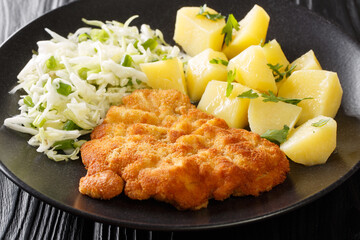Polish Breaded Pork Cutlets Kotlet Schabowy with boiled potatoes and cabbage salad close-up in a plate on a black wooden table. Horizontal - obrazy, fototapety, plakaty