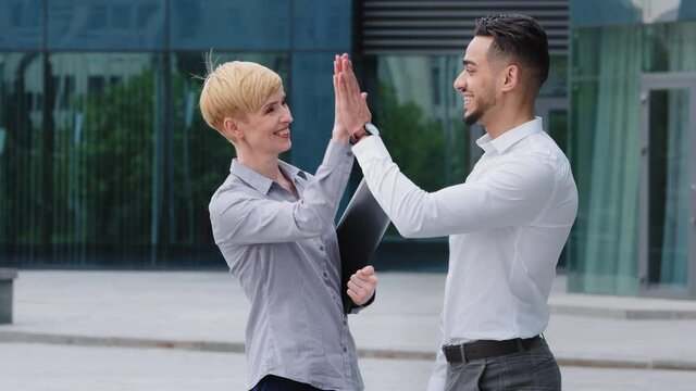 Multiracial colleagues caucasian business woman customer manager holding folder in hands stands outdoors with hispanic man investor showing hand direction to side laugh talking make high five gesture