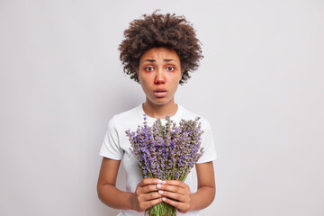 Puzzled curly haired African American woman with red eyes holds lavender bouquet has allergy...