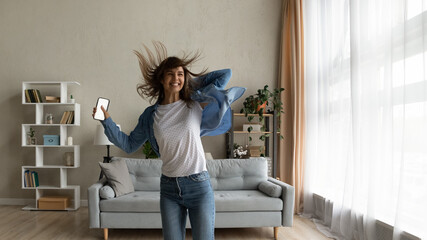 Cheery woman feels overjoyed jumps dance in living room hold smart phone with mock up white screen....