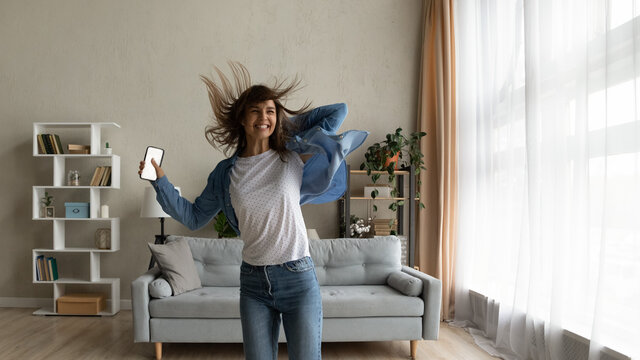 Cheery woman feels overjoyed jumps dance in living room hold smart phone with mock up white screen. Female celebrate unbelievable great news received. Fun, moment of victory, candid emotions concept