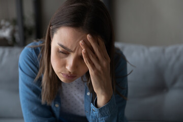 Close up of sad tired young woman sit on sofa, touch aching head having headache migraine looks...