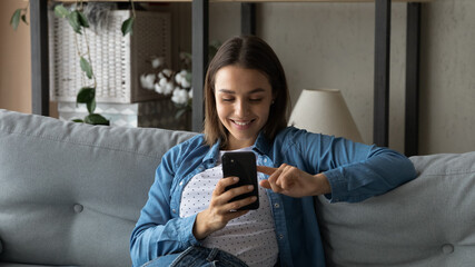 Obraz premium Lovely young woman spend time use smartphone, sit on sofa in living room hold cellphone enjoy chat in social media, search buy goods fashion clothes via e-commerce retail services application concept