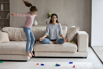 Calm young Hispanic mom sit on sofa at home meditate practice yoga keep calm relieve negative emotions. Active energetic ethnic daughter jump play near relaxed mother. Motherhood concept.
