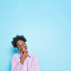 Fototapeta na wymiar Vertical shot of joyful dark skinned woman holds chin focused above with cheerful expression makes plans in mind feels happy wears purple shirt poses against blue background copy space for advert