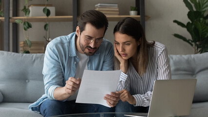 Serious pensive millennial 35s couple sit on sofa at home hold letter reading papers documents,...