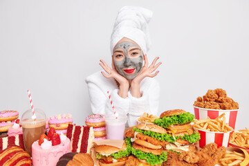 Indoor shot of pretty woman with clay mask on face red lips and manicure going to have tasty lunch sits at table full of junk food touches face gently enjoys softness of skin wears bathrobe.
